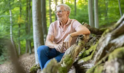 Das Bild zeigt einen Mann welcher, angelehnt an einen liegenden Baumstamm, im Wald sitzt und in die Ferne schaut.